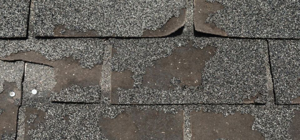 Close-up of worn asphalt roof shingles with significant granule loss, exposed underlayment, and visible nails indicating weather damage.