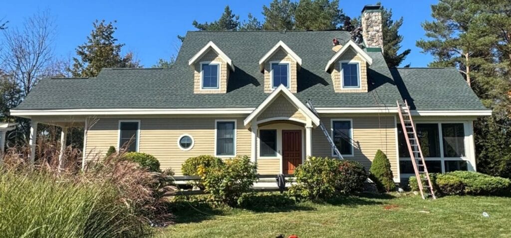 A residential home with a newly installed asphalt shingle roof, ladders set up, and roofing professionals completing work on the chimney and upper roofline.