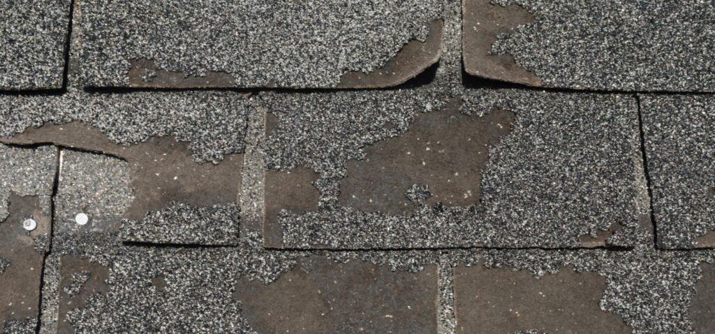 Close-up of worn asphalt roof shingles with significant granule loss, exposed underlayment, and visible nails indicating weather damage.