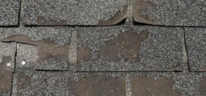 Close-up of worn asphalt roof shingles with significant granule loss, exposed underlayment, and visible nails indicating weather damage.
