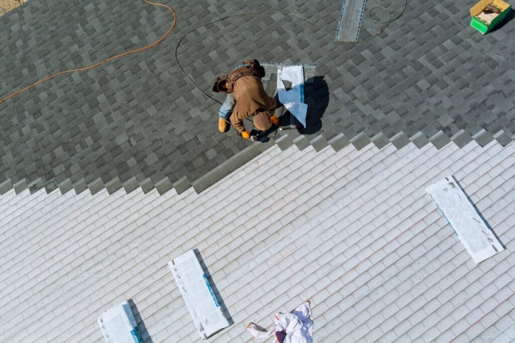 A worker installs dark asphalt shingles on a roof, transitioning from a white underlayment surface during a roofing replacement project.