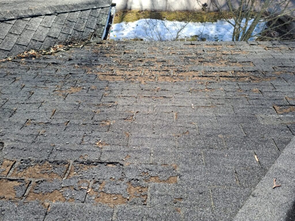 A roof with severely worn and deteriorated asphalt shingles, showing large patches where the surface granules have eroded away, exposing the underlying material.