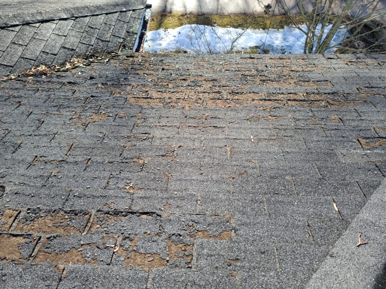 A roof with severely worn and deteriorated asphalt shingles, showing large patches where the surface granules have eroded away, exposing the underlying material.